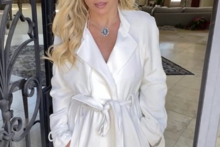 Britney Spears stand in a long white stain coat in an iron wrought doorway in the entrance to a house